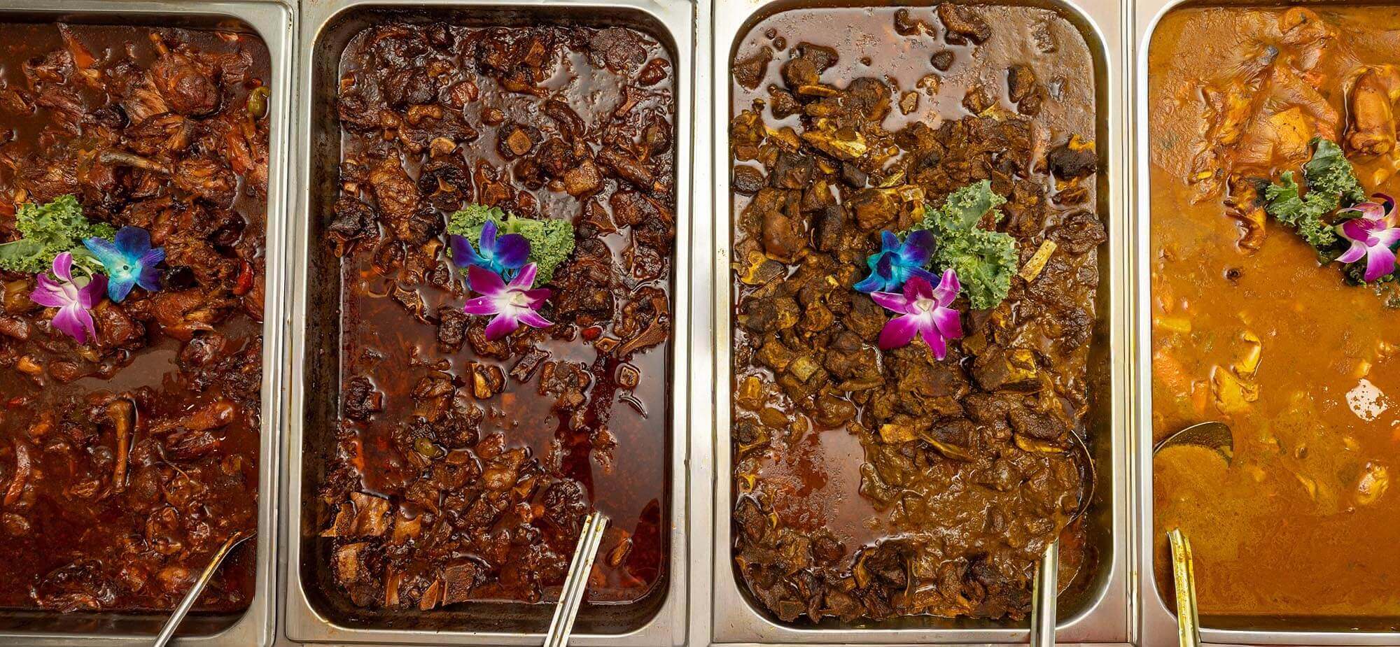 Four stainless steel trays filled with different types of cooked meat dishes in brown and reddish sauces, each garnished with green kale and purple-blue edible flowers.