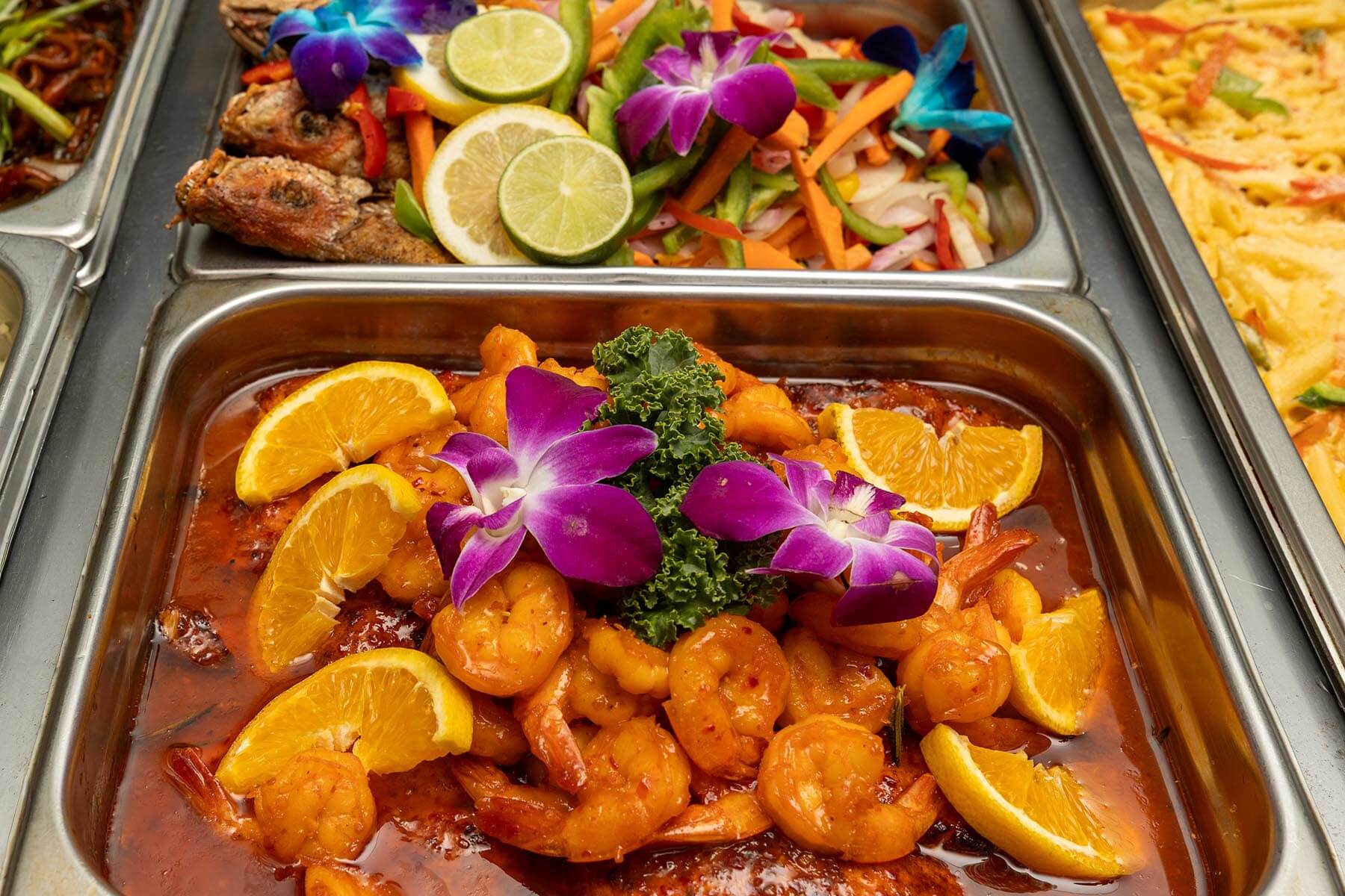 Tray of shrimp in orange sauce garnished with orange slices, purple orchids, and kale in a buffet setting.