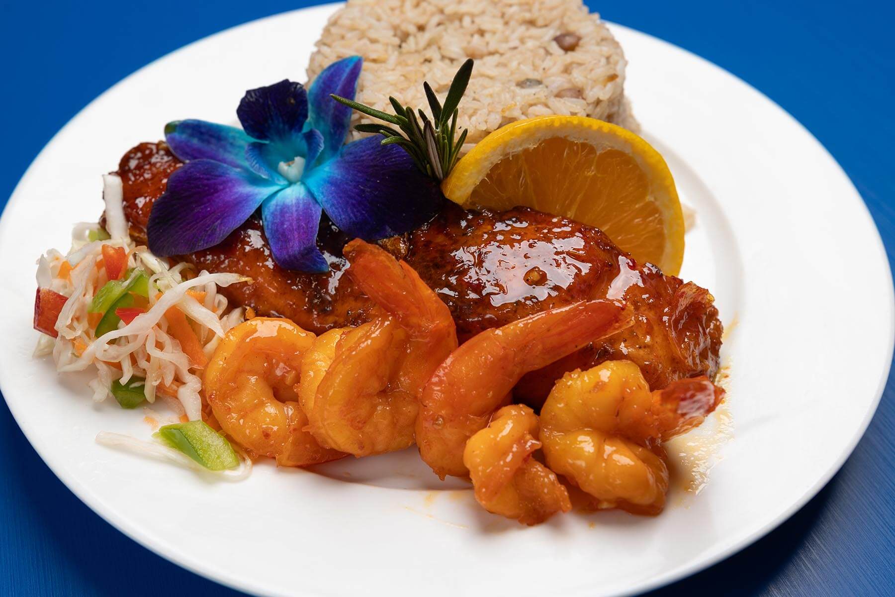 Plate with glazed shrimp and chicken, a serving of rice, coleslaw, a slice of orange, and a purple orchid garnish.