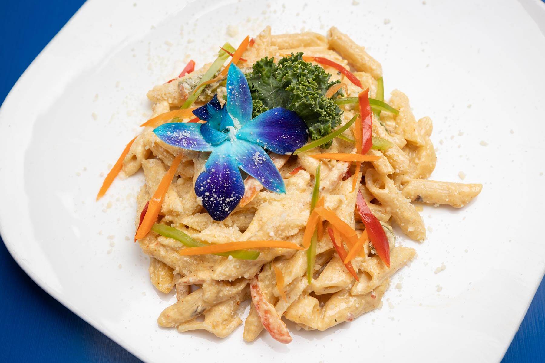Creamy penne pasta garnished with thin slices of red and green bell peppers, shredded carrot, a blue edible flower, and a kale leaf on a white plate.