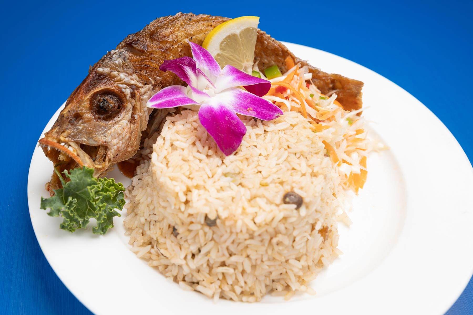 Plate with fried whole fish, mound of rice garnished with a purple orchid flower, lemon wedge, kale leaf, and shredded vegetables on a blue table.
