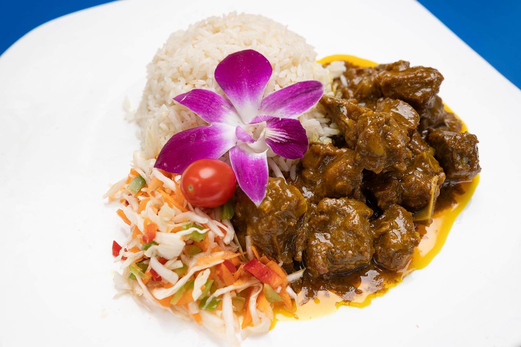 Plate with a serving of white rice topped with a purple orchid flower, brown curry beef, and a side of shredded vegetable salad with a cherry tomato.