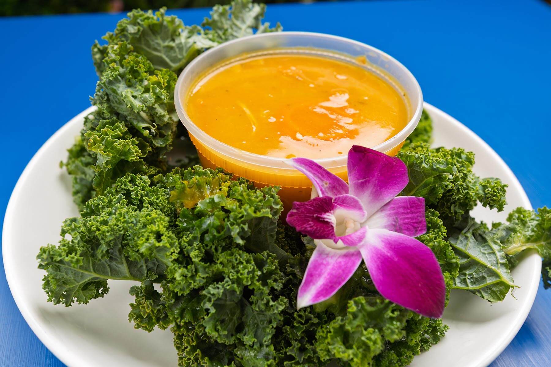 White plate with fresh curly kale leaves, a small container of orange dressing, and a purple orchid flower on a blue surface.