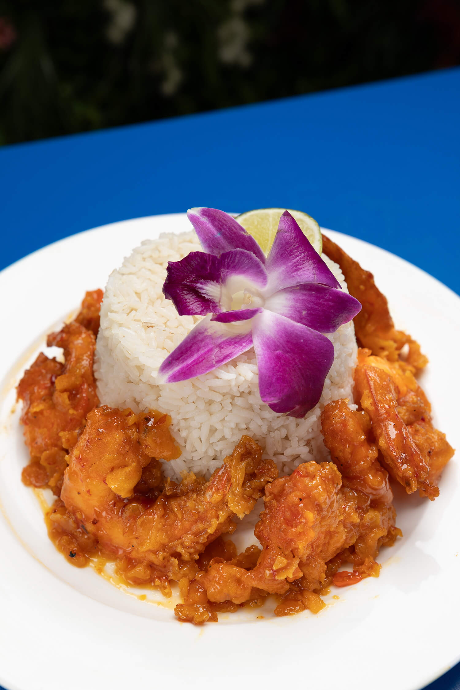 Plate of fried shrimp in sauce surrounding a mound of white rice topped with a purple orchid flower and lime wedge.