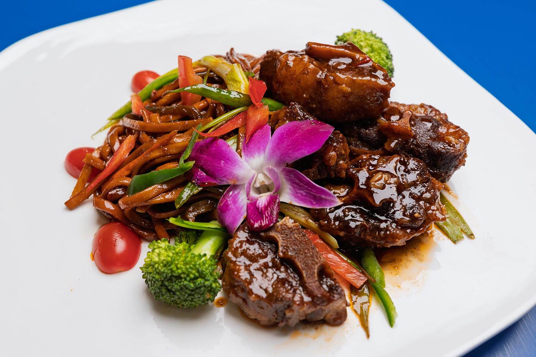 Plate with glazed braised meat ribs, stir-fried noodles with vegetables, broccoli, cherry tomatoes, and purple orchid garnish.